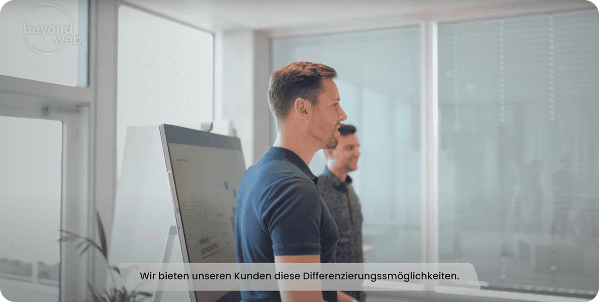 Zwei Männer stehen in einem modernen Büro neben einem Whiteboard, am unteren Bildrand ist ein deutscher Text zu sehen.