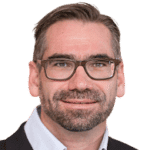 Ein Mann mit kurzen braunen Haaren, Bart und Brille trägt ein Hemd mit Kragen und eine dunkle Jacke und blickt direkt in die Kamera.