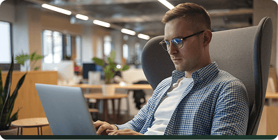 Ein Mann mit Brille und kariertem Hemd arbeitet an einem Laptop in einem modernen Büro mit offenen Sitzgelegenheiten, Pflanzen im Hintergrund und konzentriert sich auf das Schreiben von SEO-Texten.