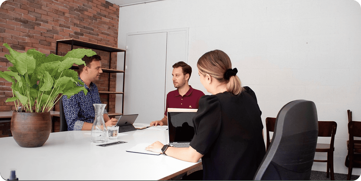 Drei Personen sitzen mit Laptops an einem weißen Tisch und unterhalten sich in einem modernen Büro mit Ziegel- und weißen Wänden. Auf dem Tisch steht eine große Topfpflanze.