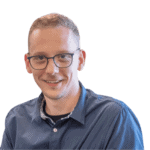 Ein Mann mit kurzen Haaren und Brille, der ein blaues Hemd mit Knöpfen trägt, lächelt, vor einem neutralen Hintergrund.