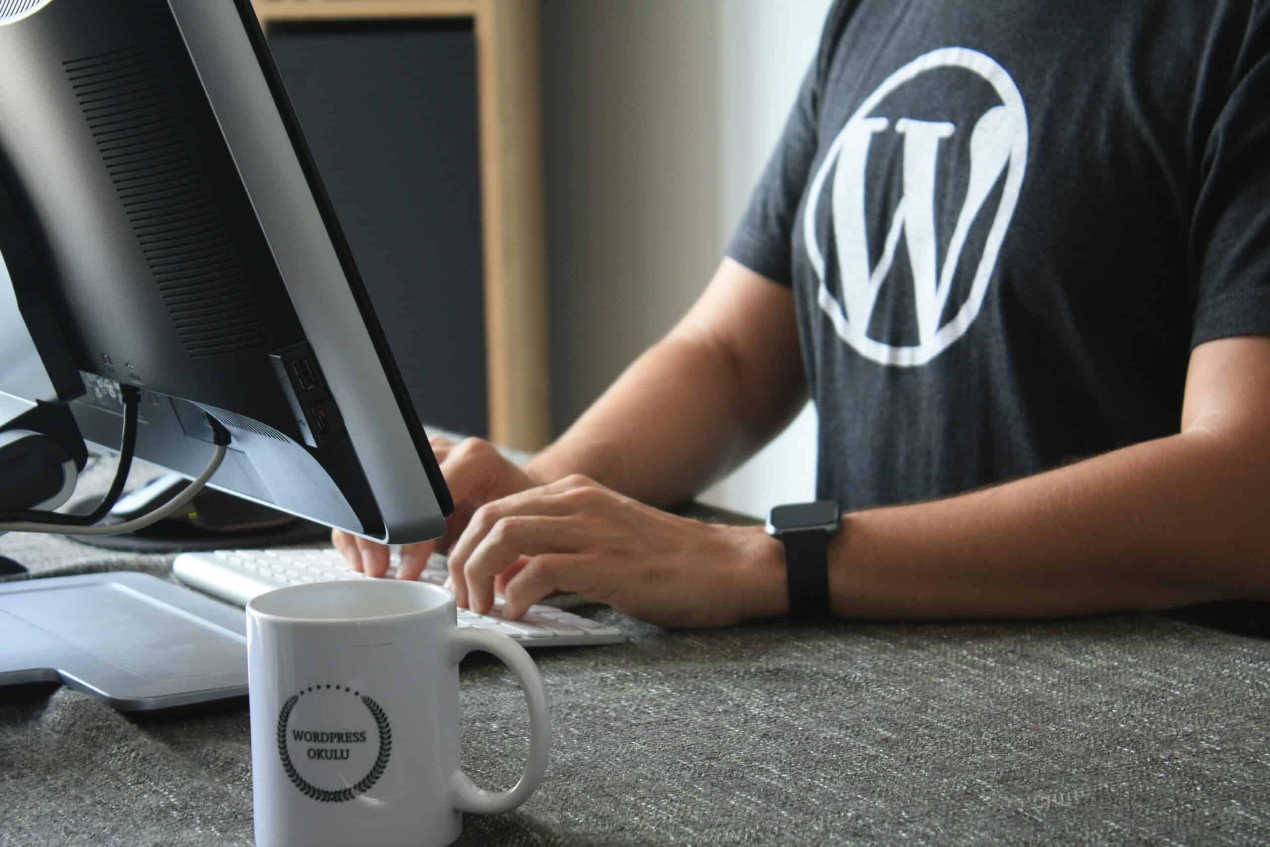Eine Person, die ein WordPress-T-Shirt trägt, tippt auf einer Tastatur an einem Schreibtisch mit einem Computermonitor und einer Tasse mit der Aufschrift „WordPress GEEKLIU“ und denkt dabei vielleicht über die Debatte Wix vs. WordPress nach.