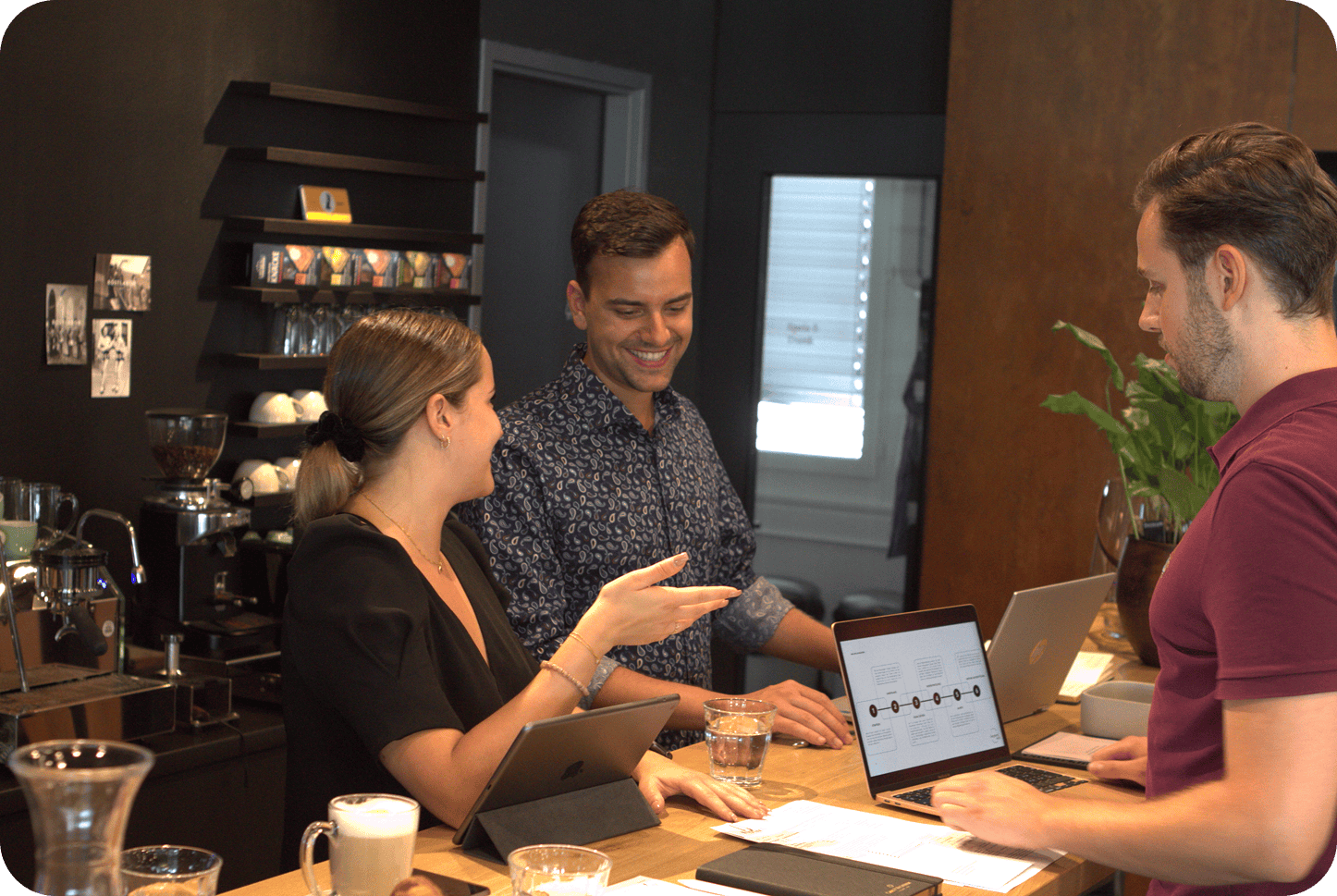 Drei Personen unterhalten sich an der Theke eines Cafés. Vor ihnen stehen Laptops und Getränke, im Hintergrund steht eine Kaffeemaschine.