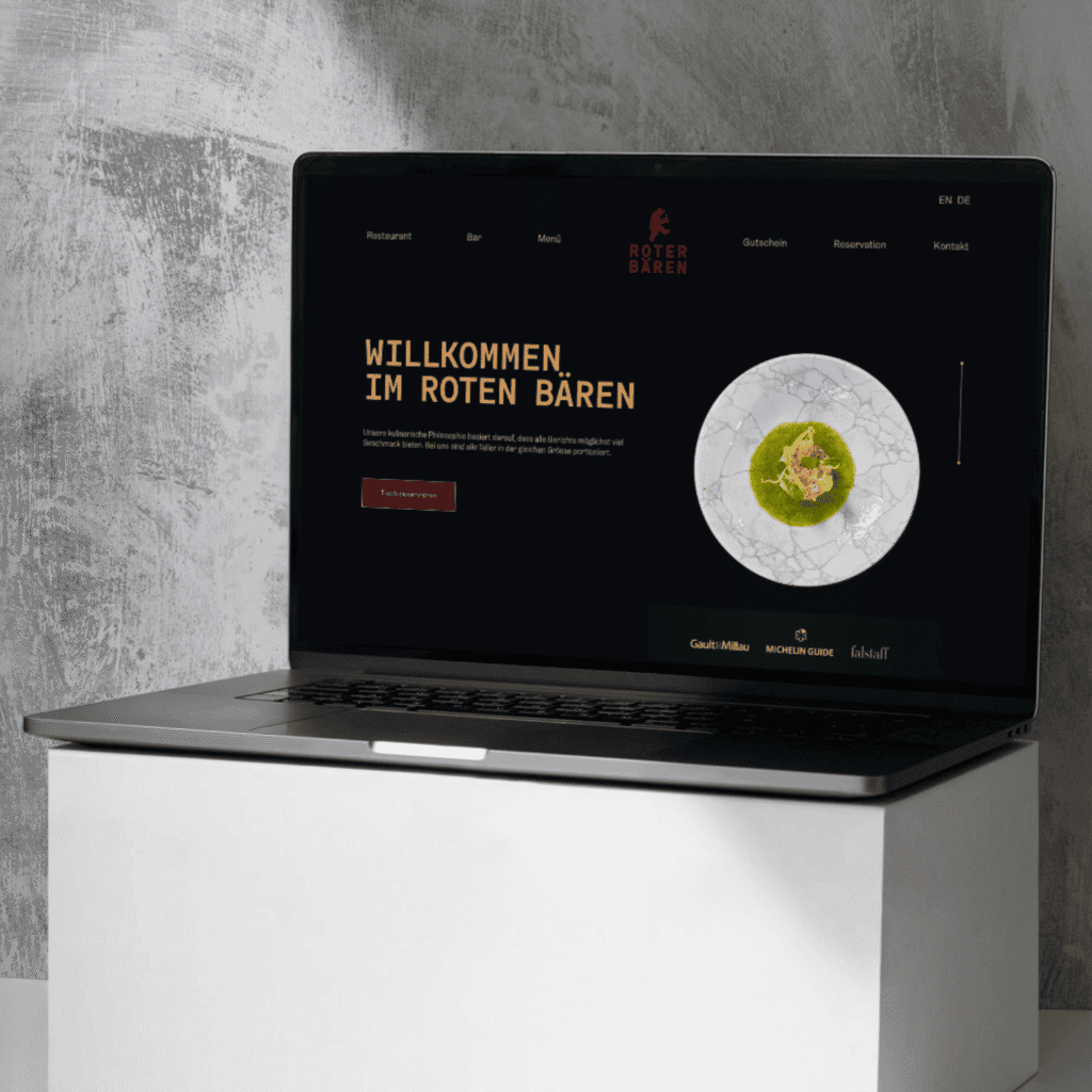 Ein Laptop, auf dem die von der Content Agentur Basel gestaltete Website des Restaurants "Roter Bären" angezeigt wird, zeigt ein dunkles Thema, einen Willkommensgruß in deutscher Sprache und ein auf dem Bildschirm abgebildetes Gericht.