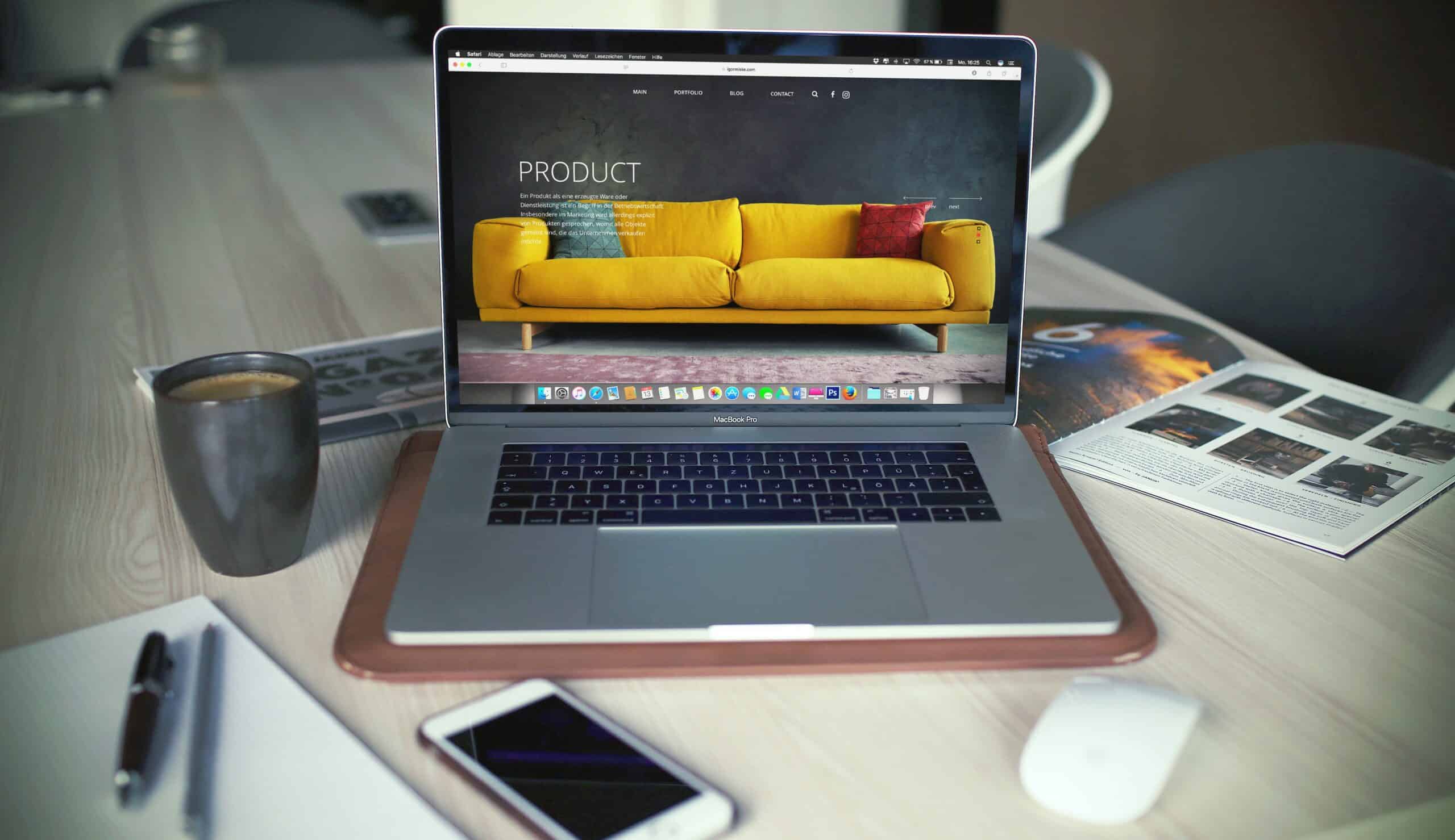 Ein Laptop mit einem gelben Sofa auf dem Bildschirm steht auf einem Schreibtisch mit einer Tasse Kaffee, einem Smartphone, einer Maus, einem Notizbuch und einer aufgeschlagenen Zeitschrift.