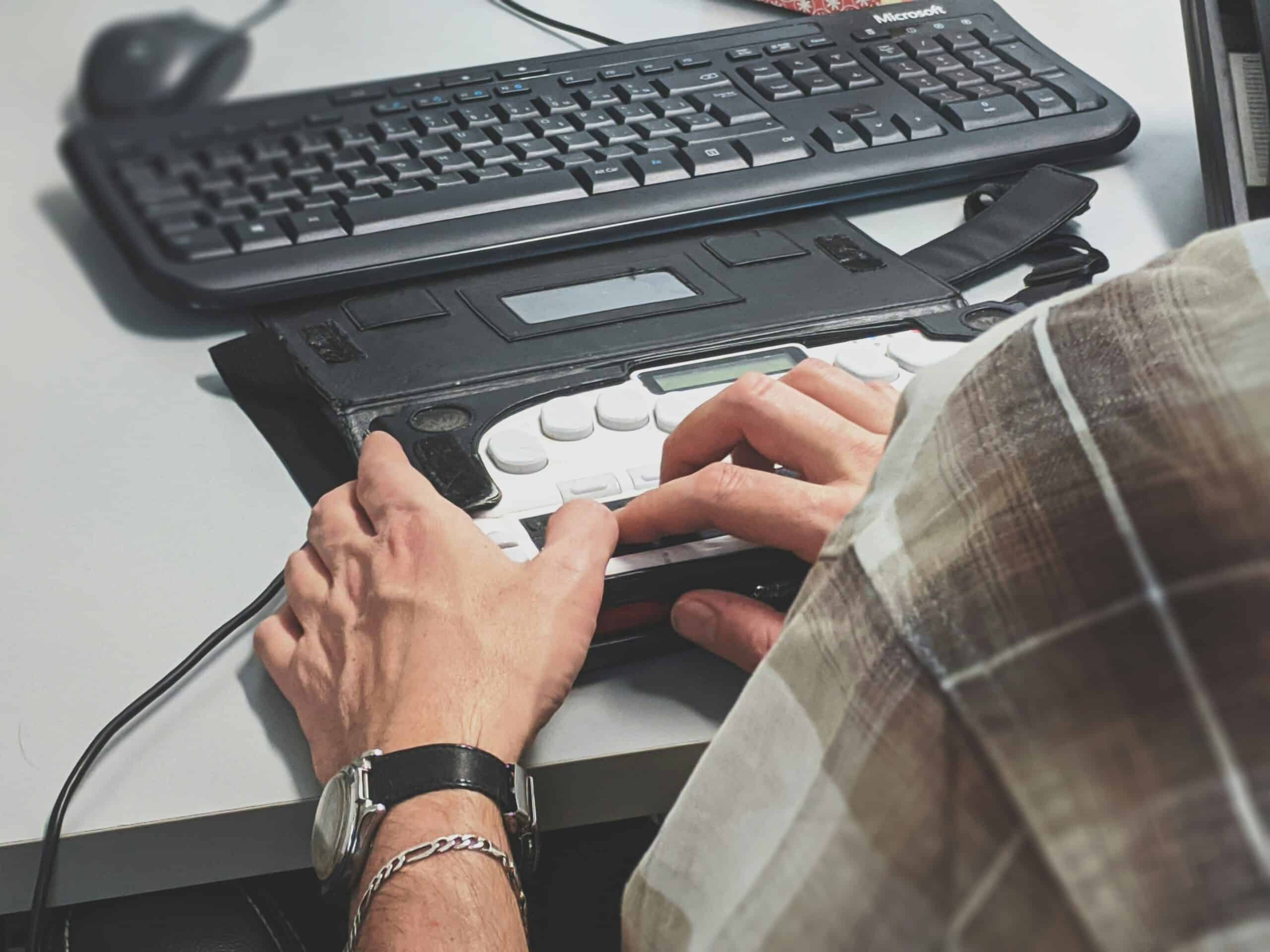 Eine Person benutzt eine Braillezeile vor einer Tastatur an einem Schreibtisch, wobei sie mit den Händen auf der Tafel liest oder tippt.