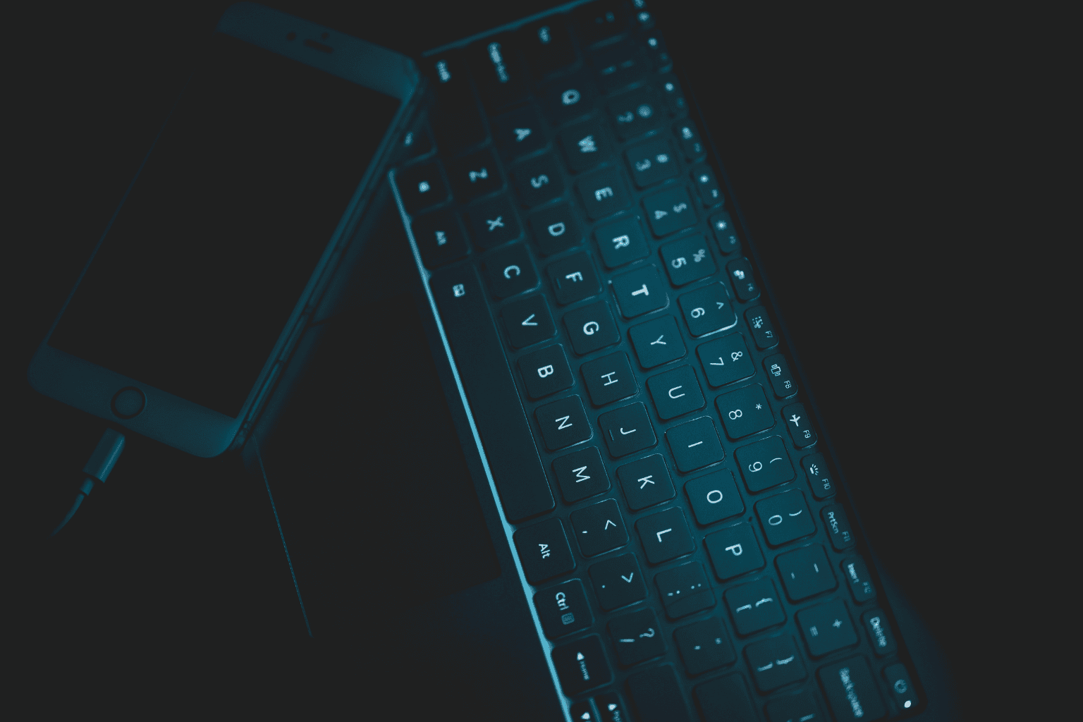 Eine Nahaufnahme einer beleuchteten Computertastatur neben einem aufladenden Smartphone in einer dunklen Umgebung.