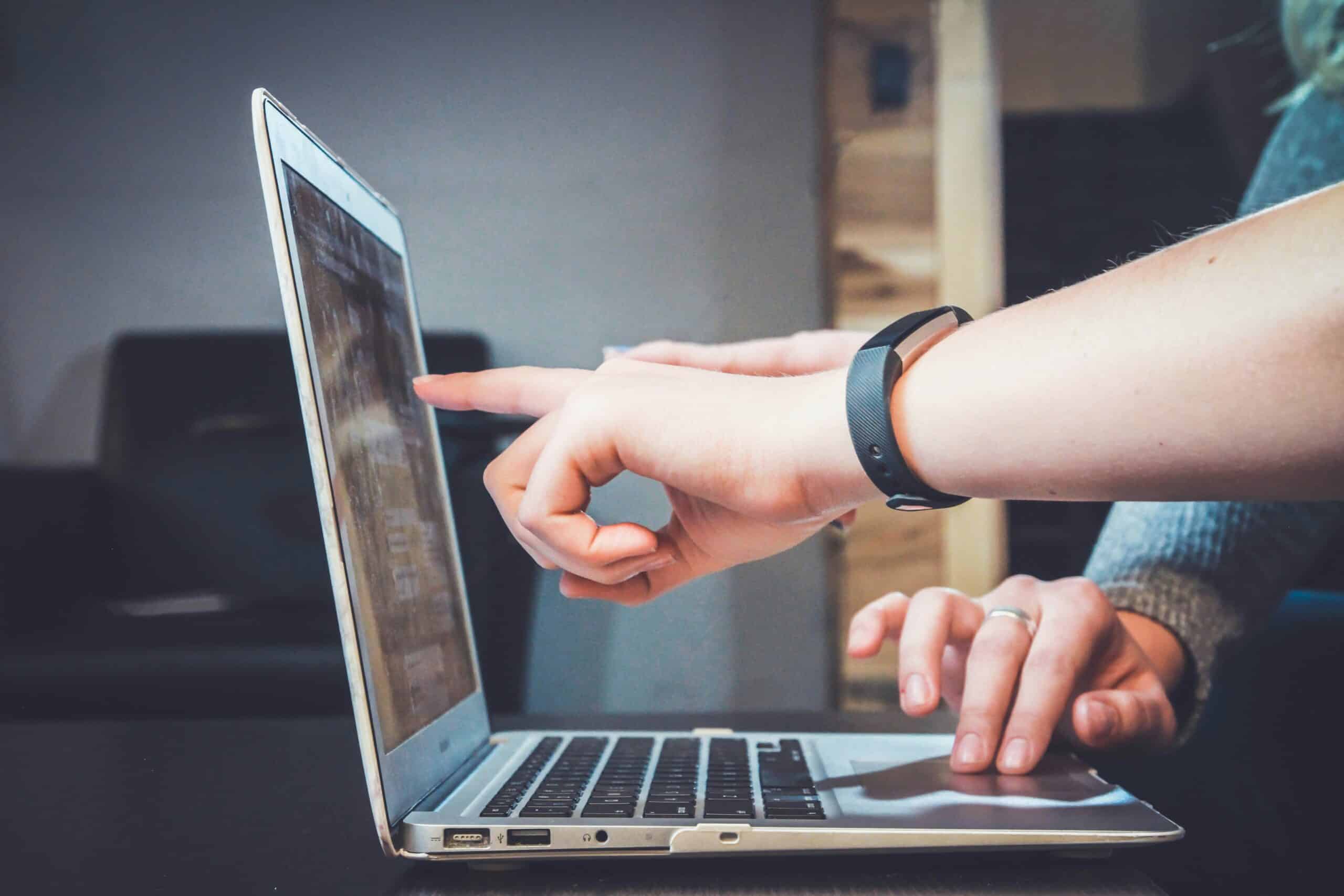 Zwei Personen interagieren mit einem Laptop; eine Person tippt, während die andere auf den Bildschirm zeigt und möglicherweise gemeinsam etwas diskutiert oder bespricht.