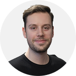 Ein junger Mann mit heller Haut, kurzen braunen Haaren und einem gestutzten Bart trägt ein schwarzes Hemd und lächelt in die Kamera vor einem schlichten weißen Hintergrund.