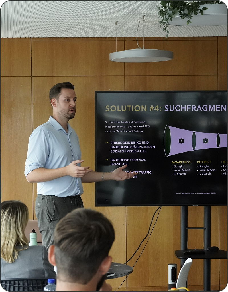 Ein Mann steht auf und präsentiert dem Publikum Informationen auf einem Bildschirm; auf dem Bildschirm erscheint eine Folie mit dem Titel "LÖSUNG #4: SUCHFRAGMENTE".
