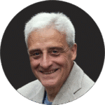 Ein älterer Mann mit kurzen weißen Haaren und einer hellgrauen Jacke lächelt in die Kamera vor einem dunklen runden Hintergrund.