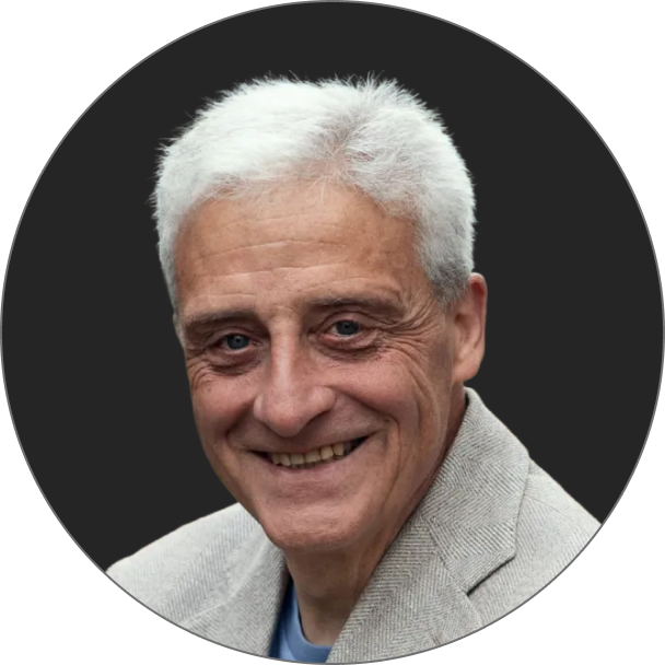 Ein älterer Mann mit kurzen weißen Haaren und einer hellgrauen Jacke lächelt in die Kamera vor einem dunklen runden Hintergrund.