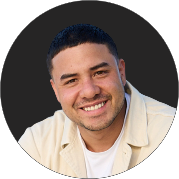 Ein Mann mit kurzen dunklen Haaren und Gesichtsbehaarung lächelt in die Kamera. Er trägt eine helle Jacke über einem weißen Hemd und steht vor einem dunklen, runden Hintergrund.