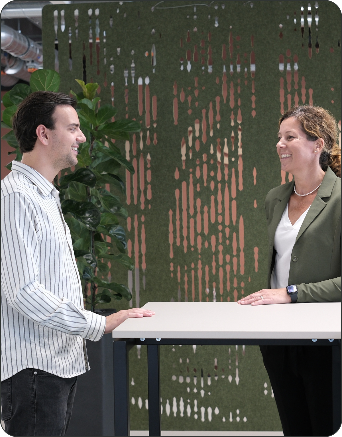 Zwei Personen stehen und unterhalten sich an einem hohen Tisch in einem modernen Büroraum mit Grünpflanzen und einem dekorativen Raumteiler im Hintergrund.