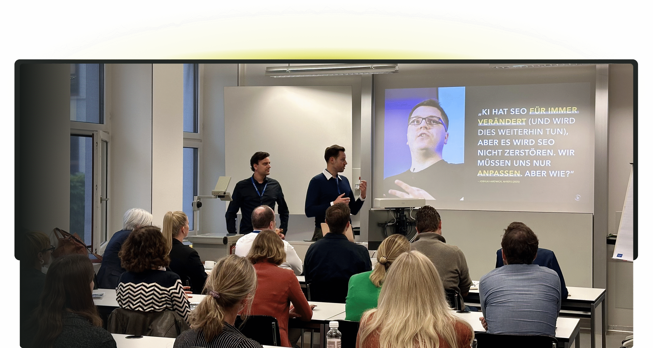 Zwei Männer stehen und präsentieren vor einem sitzenden Publikum in einem Klassenzimmer während eines SEO-Workshops; eine Folie mit deutschem Text über SEO wird auf die Leinwand hinter ihnen projiziert.