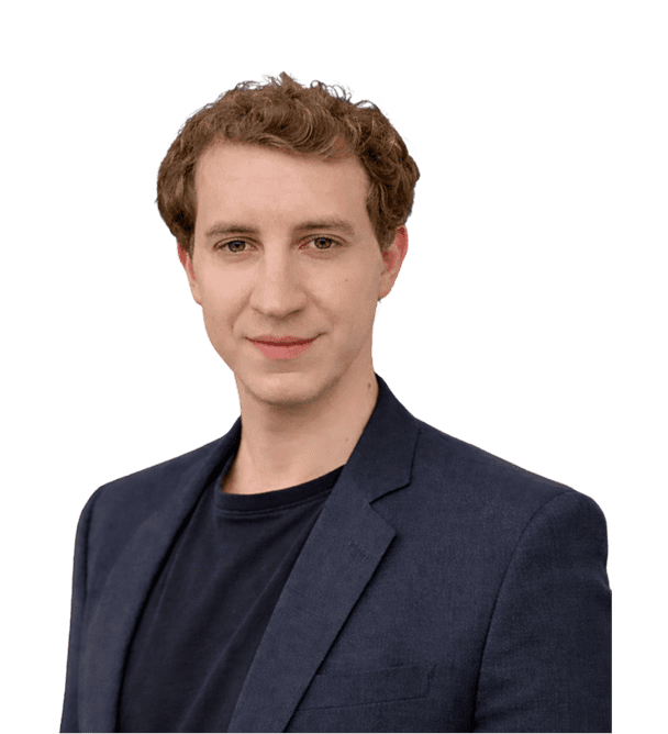 Ein Mann mit kurzen lockigen Haaren, der einen dunklen Blazer und ein schwarzes Hemd trägt, steht vor einem schlichten weißen Hintergrund.