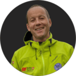 Mann, der eine hellgrüne Outdoor-Jacke mit Logos trägt, lächelt und vor einem schlichten dunklen Hintergrund posiert.