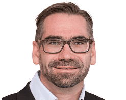 Mann mit kurzen dunklen Haaren, Bart und Brille, der eine Anzugsjacke und ein hellblaues Hemd trägt und vor einem schlichten weißen Hintergrund posiert - ideal für Profile im Zusammenhang mit Websites für Softwareunternehmen.