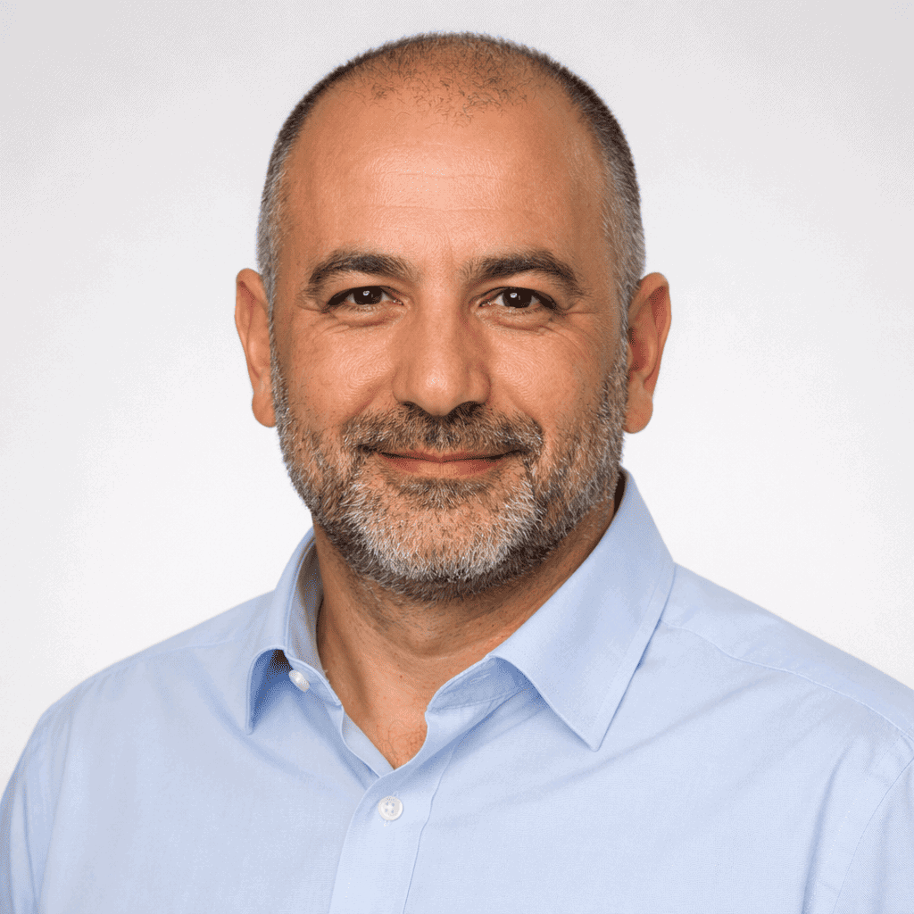 Ein Mann mittleren Alters mit rasiertem Kopf und grauem Bart, der ein hellblaues Hemd mit Kragen trägt, lächelt in die Kamera vor einem schlichten Hintergrund.